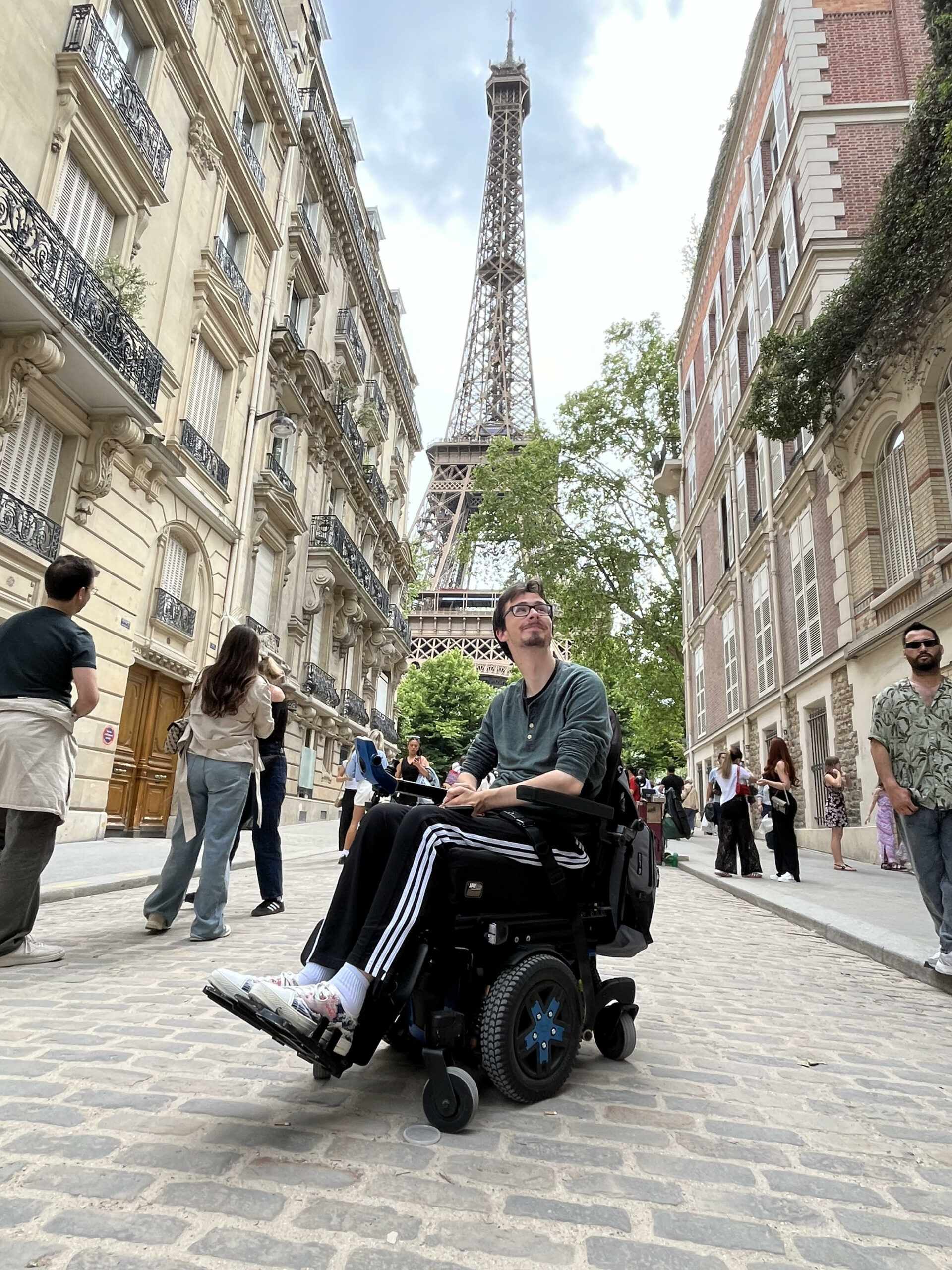 Dexter in power wheelchair on cobblestone street with buildings and pedestrians on either side with view of Eiffel Tower behind him