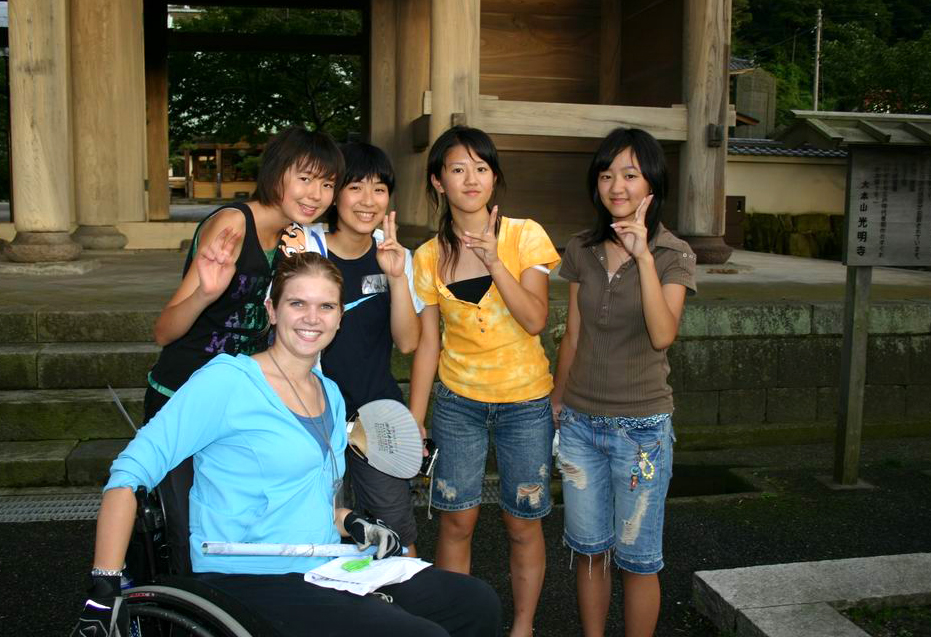 (a woman wheelchair rider with some other women in an Asian country who are standing next to her and smiling and gesturing