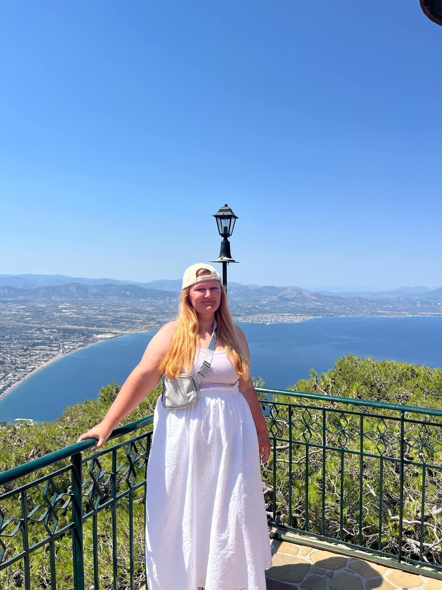 Johna in Corinth, Greece standing at the top of the monestery overlooking the sea. wearing a backwards hat, pink tank and white skirt