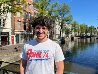 Nathaniel stands outdoors in front of a canal lined by trees and buildings. He wears a Bowie 74 world tour t-shirt and smiles