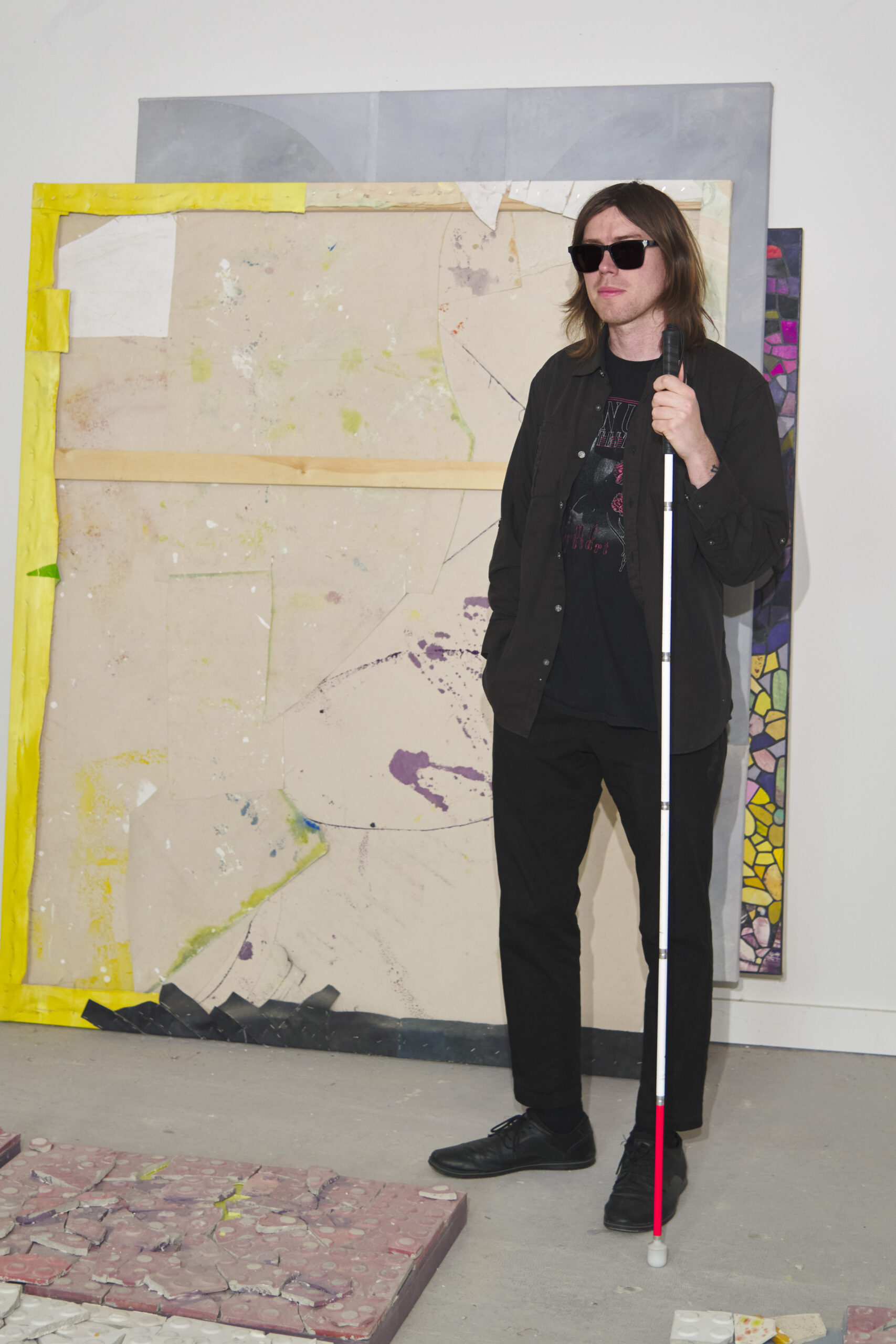 Stephen stands in front of a large canvas. He wears all black, dark sunglasses and holds a white cane.