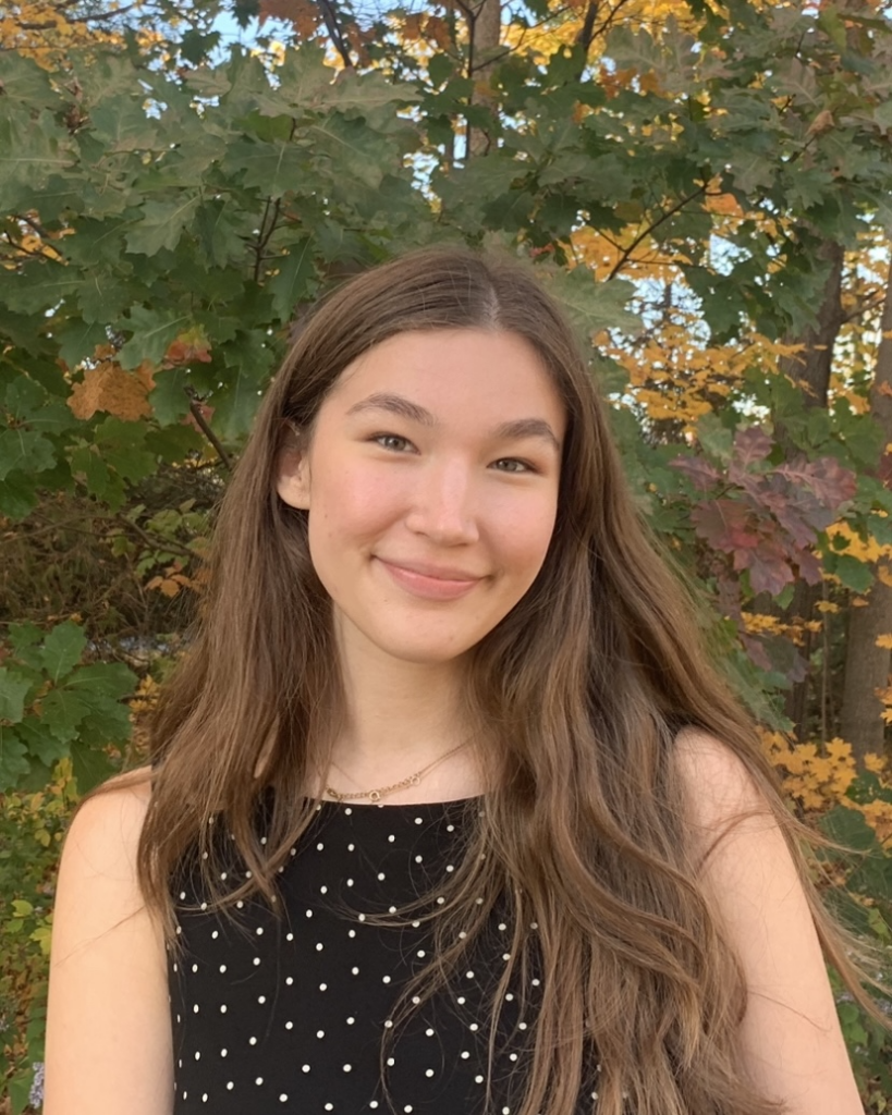 Aubrianna, a young biracial Japanese and White disabled woman, smiles outside in front of fall leaves. She has long brown hair and is wearing a black dress with white polka dots.