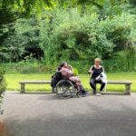 GDDI 2022 cropped A black woman using a wheelchair talking with a lighter skinned woman outside surrounded lush nature.