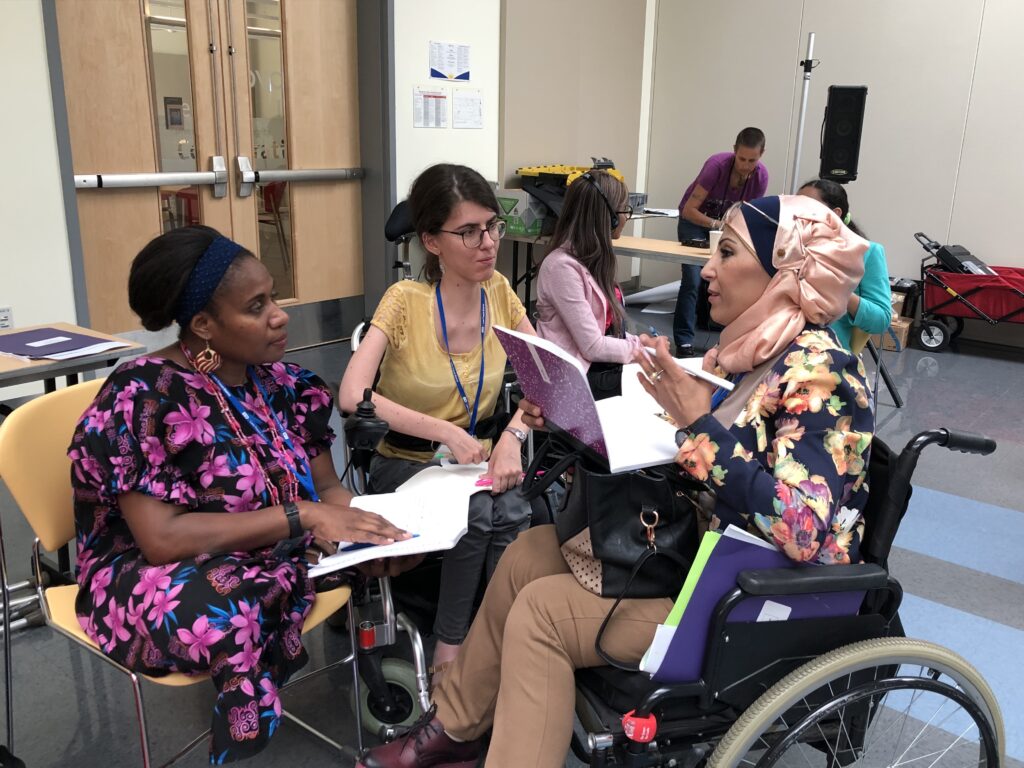 Three women in a conference room gather around a small table. Two women are seated in wheelchairs. One woman wears a headscarf. The three women are different ethnicities/races and wear colorful blouses or dresses.