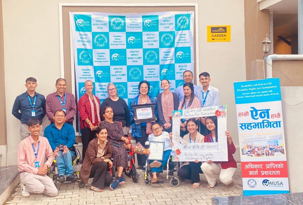 Group photo of CIL-P and MIUSA staff in front of a banner with both organizations' logos. Two staff hold a sign that reads "Our laws, our participation, paving the way for the attainment of our rights!"