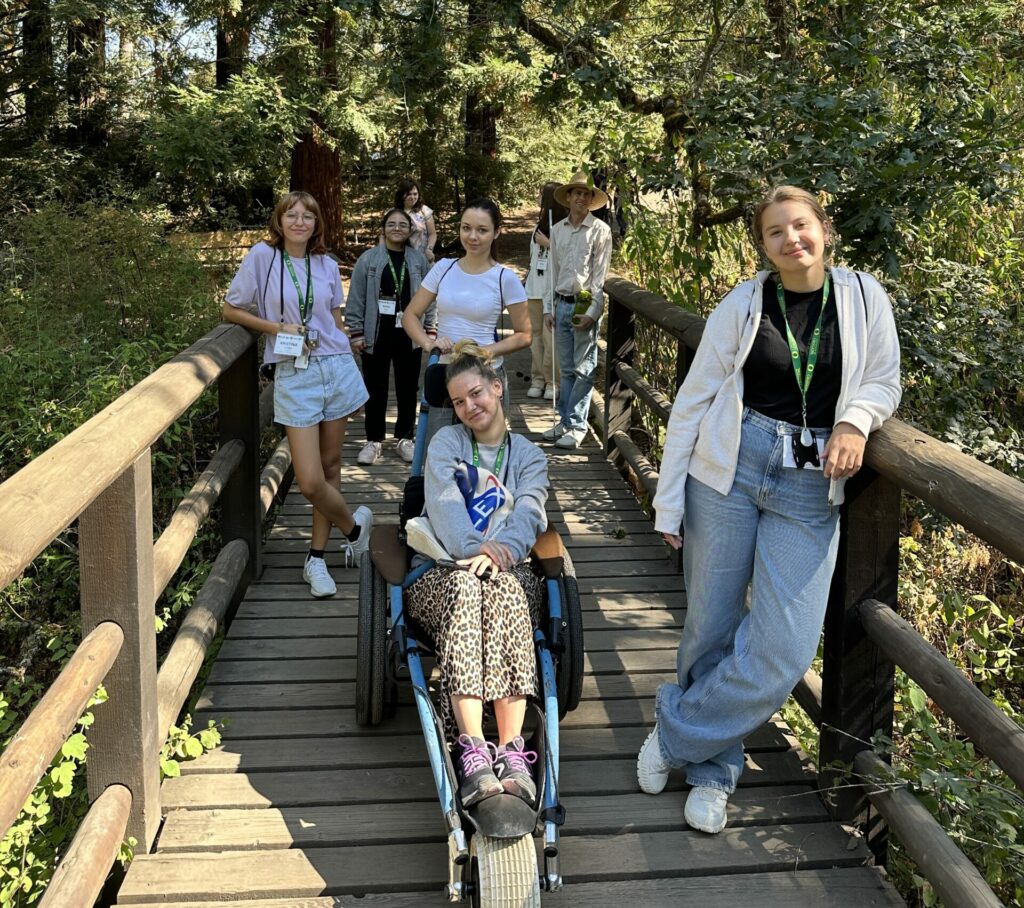 Students standing on a wooden bridge smiling with trees in the background. One student rides in a wheelchair with a long front wheel and two larger back wheels.