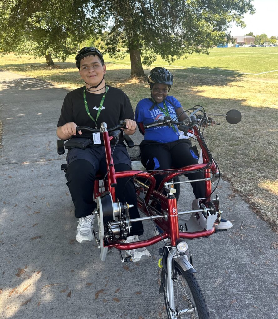 Two students ride a side-by-side tandem tricycle together on a bike path.