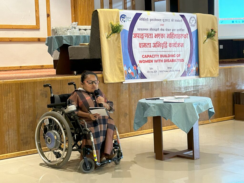 Anjana, a wheelchair rider, speaks into a microphone. Behind her is a banner with MIUSA logo and CILP logo and text in Nepali.