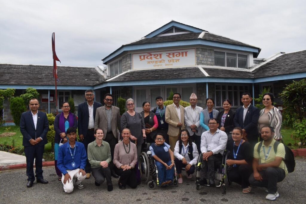 Group photo of approximately 20 people, including two wheelchair riders, posing in front of a government building with the Nepal flag behind them.