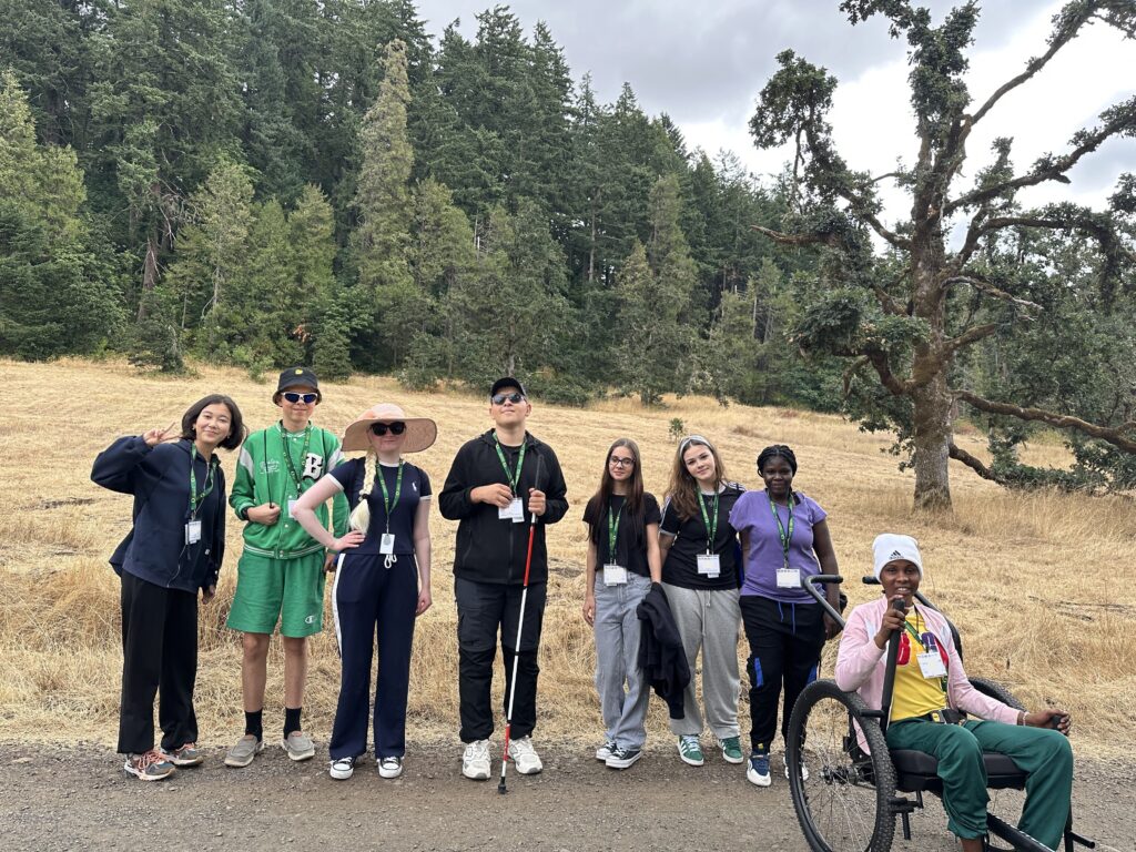 Group of students including one riding a wheelchair and one holding a white cane smiling on a gravel trail with trees behind them. 