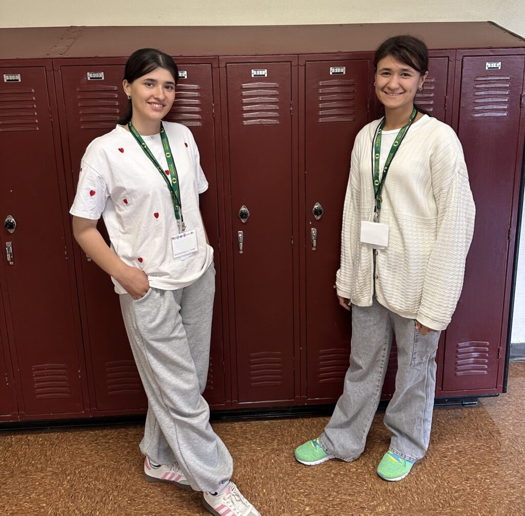 Two students smile and stand in front of a row of lockers.