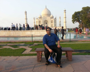Qazi Fazli Azeem, a middle-aged man with dark hair and skin dressed in blue button-down shirt and black slacks, rests on a bench in a courtyard. A bit farther behind him the ivory-colored Taj Mahal is visible, surrounded by visitors.
