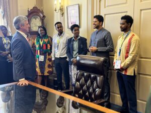 A group of seven people - black women and men in their 20s and 30s who are dressed formally in blazers or clothing in colorful African prints - stand side by side inside an office next to large doors, near a large desk with polished leather chair. One young man smiles in discussion with a man, who is white and in his 50s or 60s with gray hair and wearing a blue suit, standing with a hand resting on the desk as he faces the group.