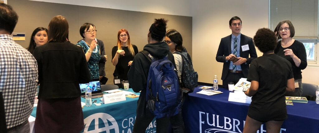 A diverse group of people at Gallaudet University's Go Global Fair in D.C. There are two information booths with opportunity representatives standing behind and Deaf students in front of the booths communicating. There appear to be sign language interpreters providing translation services for the people communicating.
