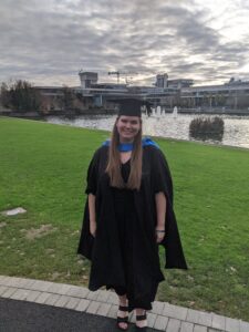 Lindsey stands outdoors wearing black graduation cap and gown, with a blue stole around her neck. Behind her is a wide gray building with reflecting pool/water fountain and green lawn. The sun shines through overcast sky.