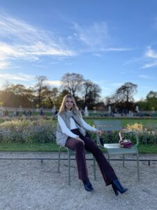 A white woman with blonde hair seated on a chair in front of a garden.