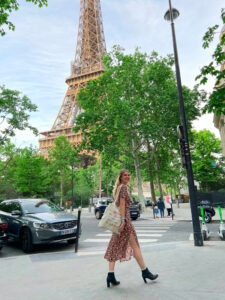 Kristen, a white woman with blonde hair, stands in front of the Eiffel Tower, mid stride, holding a bag.