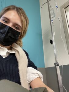 A white woman with blonde hair wearing a black facial mask and connected to an IV.