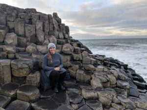 Lindsey, a white woman in her 20s or 30s, sits on a rock formation next to a calm sea and soft gray sky. She wears a long blue sweater and hat and smiles at the camera.