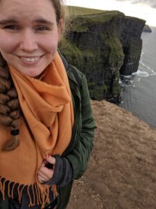 Selfie of Lindsey smiling into the camera, wearing a large orange scarf and green sweater and hair braid draped over one shoulder. In the distance behind her is a dark sea from which high green-capped cliffs emerge.