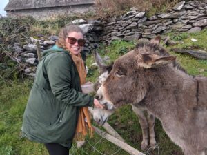 Lindsey, wearing a coat and scarf, leans over a low fence, holding a container of food in one hand, and allowing one of two gray-brown donkeys to eat from her other hand. Lindsey looks over her shoulder to smile at the camera.