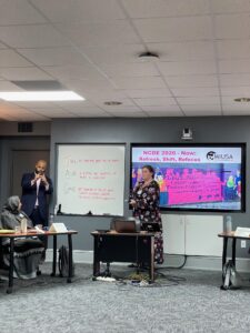 Lindsey speaks into a microphone in a conference room in front of a screen displaying a colorful presentation and MIUSA logo. A sign language interpreter nearby stands, signing as Lindsey speaks. A woman wearing a gray head scarf and dress listens, seated at a small table.