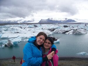 Two young women in their 20s stand, one with arms wrapped around the other’s shoulders while smiling at camera. Both women have light skin and dark hair and wear coats (one blue, the other pink) for hiking in very cold weather. The landscape behind them is comprised of grey-blue sea, ice-blue glaciers, and slate blue mountains and white-grey sky.