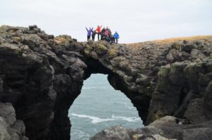 A small group of people, appearing tiny due to being viewed from far away, standing atop a massive bridge-shaped rock formation through which the sea is visible. The people wear colorful jackets and have arms outstretched, although their faces are too far away to be distinguishable.