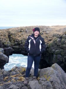 Bethany stands on and in front of a cluster of dark moss-covered rocks. She faces the camera and wears hiking boots, jeans, and cold-weather jacket. She has light skin and her hair is covered by a knit hat.