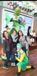 Three women in their late teens or early 20s with dark hair and light or olive skin, stand together smiling at the camera, dressed for cold weather and carrying bags. In front and behind them are circus performers dressed in bright green and colorful makeup - one standing on stilts, and the other carrying juggling pins.