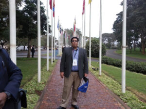 Fazli, wearing a suit and a badge with lanyard, stands outdoors on a brick walkway flanked by dozens of flag poles.