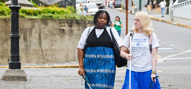 A black blind woman and a white blind woman walking down the street together. Both woman are using white canes.