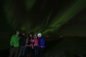 Five people huddled together, bundled up in ski jackets, scarves, and hats. It’s nighttime and behind them are the northern lights, which manifest as bright green streaks against the black sky.