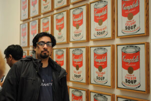 Fazli, wearing dark t-shirt and jacket, gazes at the camera with a neutral expression. Behind him is a wall displaying several individual framed prints of Andy Warhol’s Campbell soup can art.