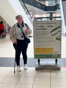 Lindsey stands smiling at camera in a well-lit building, one hand resting on a cane, the other hand giving thumbs up. Around her neck is a green lanyard with sunflower badge. Next to her is a sign that reads “Not every disability is visible - some are hidden.”