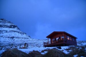A small wooden cabin surrounded by a deck emits warm light through the windows, in contrast to the bright white snow-covered hills surrounding it, and a sky that is blue but also dark, as if at dawn or dusk.