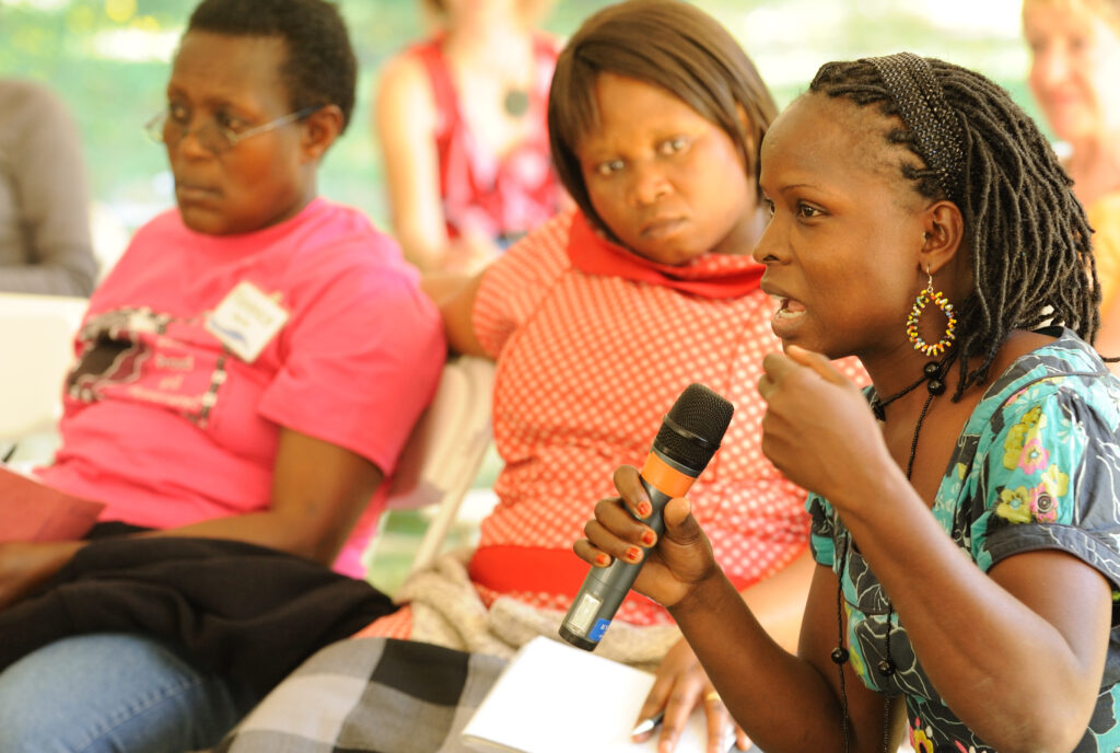 Three women sitting outside. One woman holds a microphone while speaking passionately.