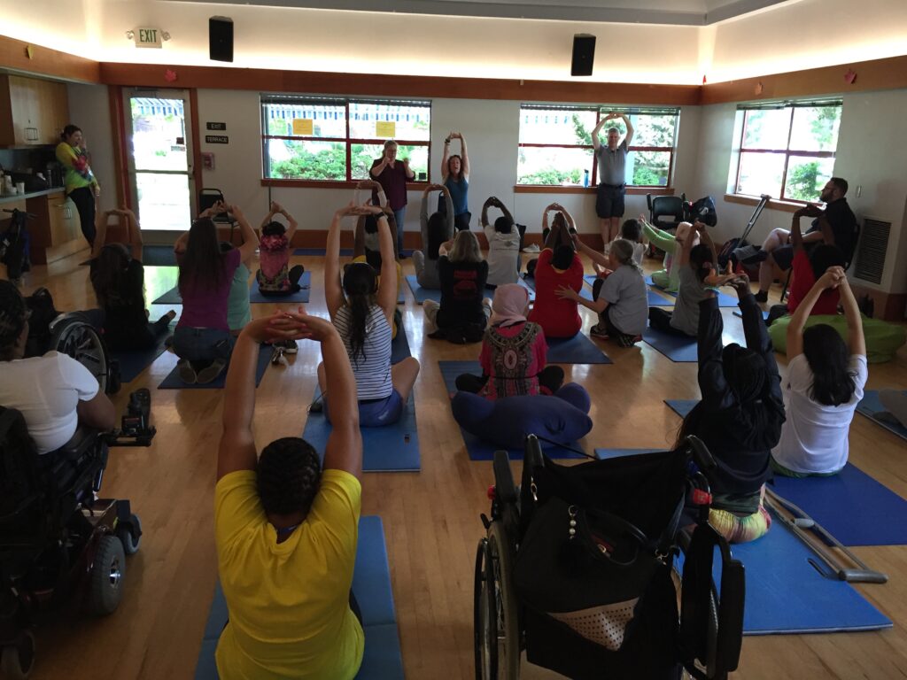 View from the back of a room of women sitting on yoga mats with their arms raised overhead. An empty wheelchair is visible next to one woman.