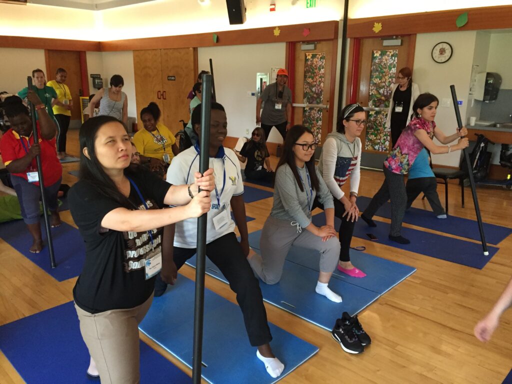 Group of women in a yoga class doing a standing lunge. A few women hold a vertical bar for balance.