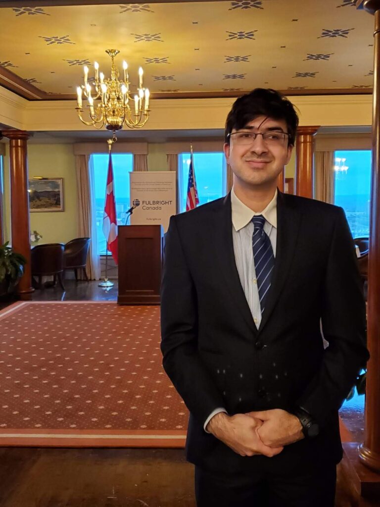 Omar with dark brown hair standing and wearing glasses, a white shirt, blue striped tie, and suit set with hands clasped. Background is a formal meeting room in the Rideau Club building in the city of Ottawa with a chandelier hanging from the ceiling. On the ground level, there is the Canada flag, the U.S. Canada, a podium, and a banner saying Fulbright Canada.