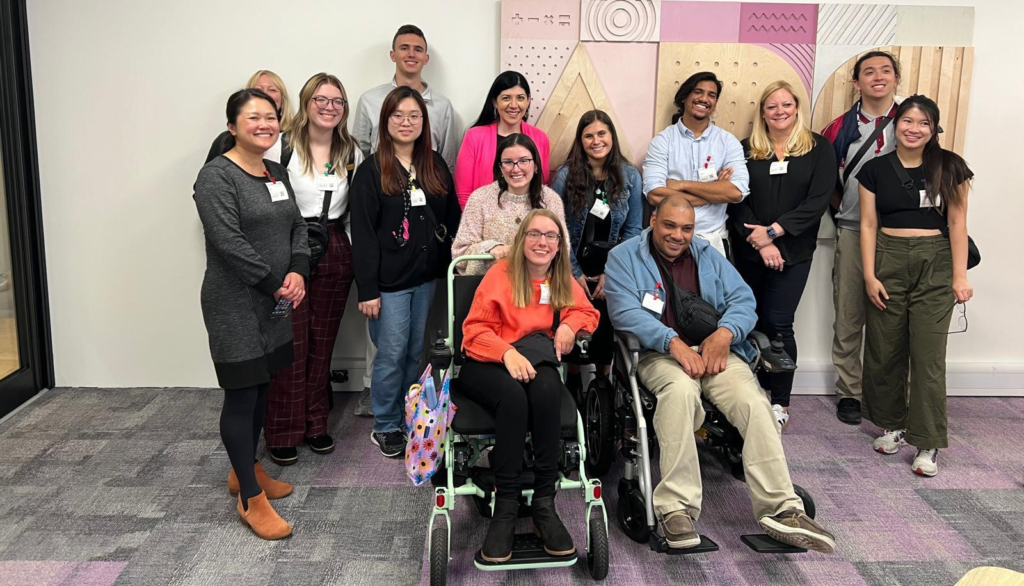 A diverse group of 15 people, including two in wheelchairs, smiling and posing together in front of a wall with abstract geometric patterns.