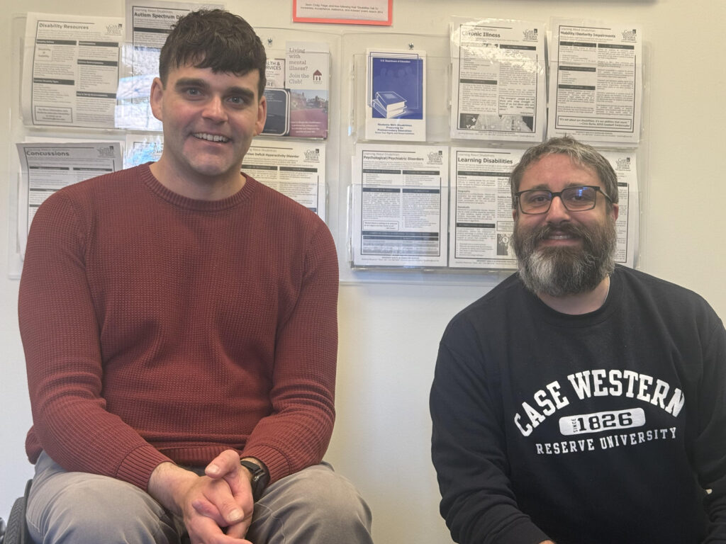 Two men sitting in front of a wall with informational posters, one wearing a red sweater and the other a "Case Western Reserve University" sweatshirt.