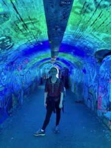 A young woman stands smiling in a graffiti-covered tunnel lit with vibrant blue and green lights. She wears a patterned blouse, leggings, hiking shoes, and a backpack. The narrow brick tunnel is filled with colorful street art, creating a glowing, surreal atmosphere.