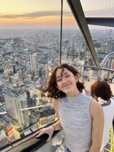 A young woman smiles at the camera while standing on an outdoor observation deck high above a sprawling cityscape at sunset. She wears a sleeveless gray top and has short, wavy brown hair. Behind her, the skyline of Tokyo stretches into the distance, with tall buildings and streets visible far below, and warm orange and pink hues coloring the sky.