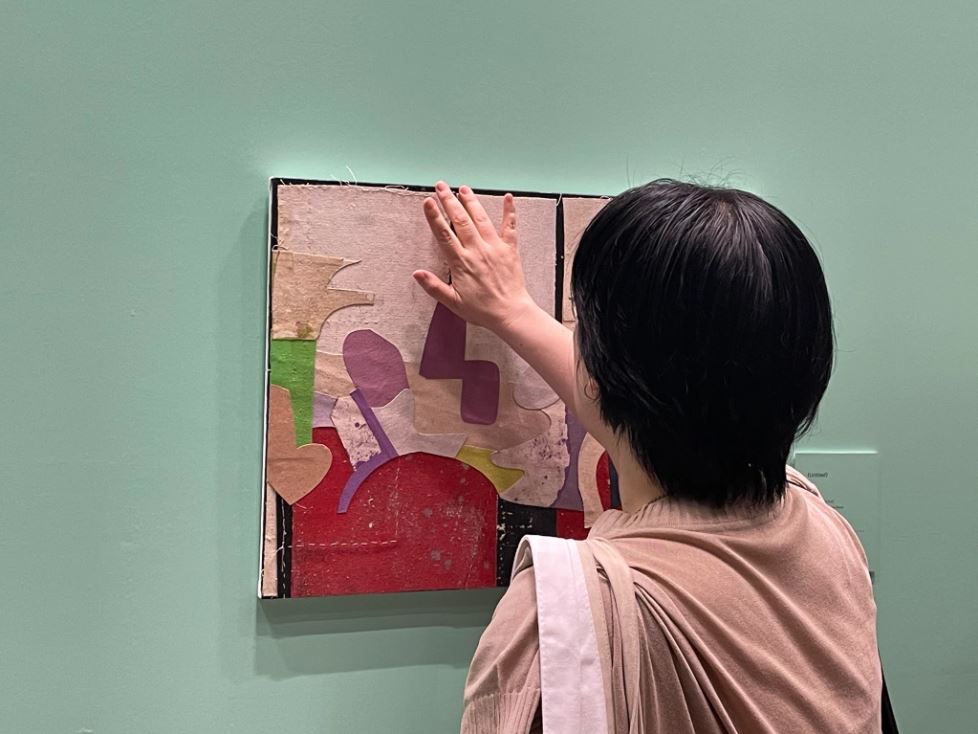 Inside an art gallery, a woman with fair skin and short black hair runs her hand along a small rectangular artwork hanging on a seafoam colored wall. The piece is tactile and includes a collage layering colorful shapes cut from fabric or other media.