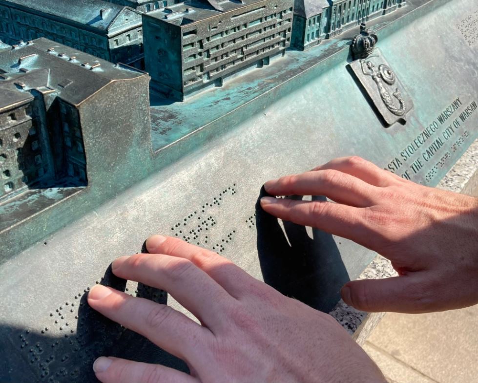 Close-up of two hands touching the braille of a bronze plaque. Next to the braille is some raised text in Polish and in English. Above the plaque is a miniature 3-D representation of various buildings and streets.