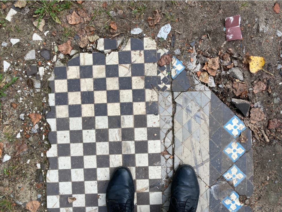 View from above looking down toward two black shoes - presumably Stephen's feet - standing atop broken slabs of checker patterned tile on dirt, grass, and dry leaves