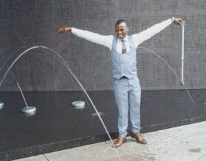 A young black man wearing suit vest stands in front of a grey fountain with arms outstretched and wide smile