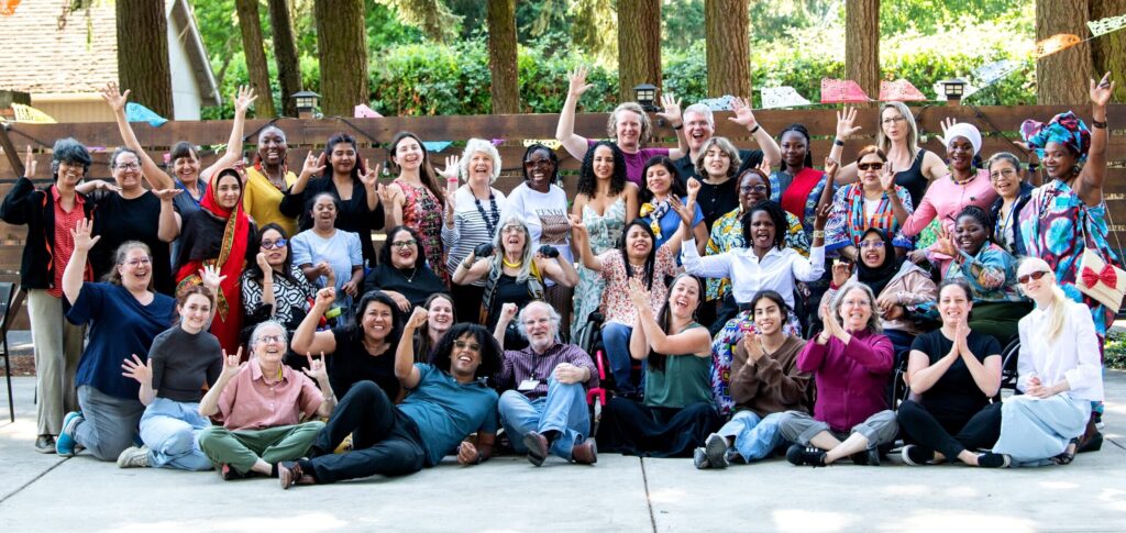 Large group of women of diverse races and disabilities, gathered in a group outdoors under brightly colored paper flags.