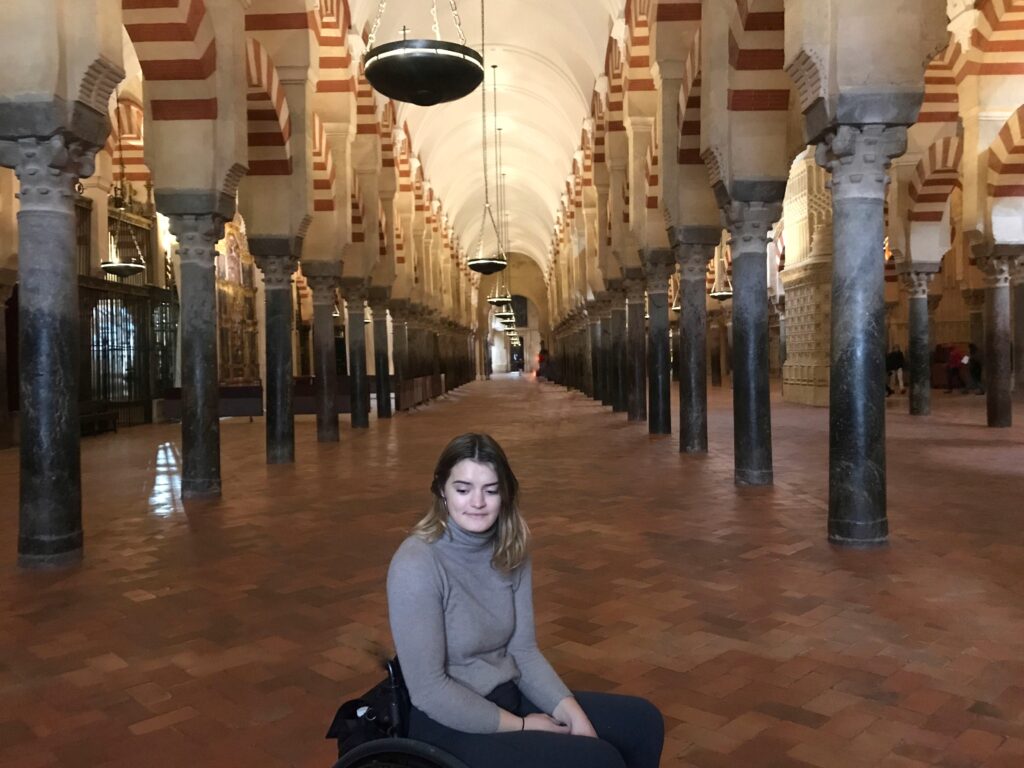A young woman in a wheelchair sits on the tiled floor of a large hall with many striped arches and pillars.